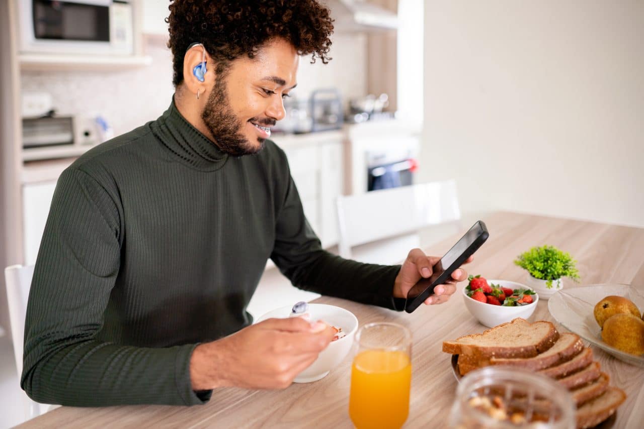 Smiling young man with hearing aids looking at his phone over breakfast.